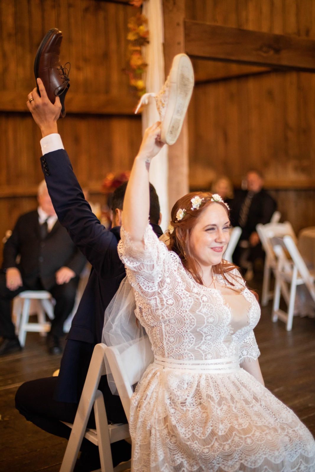 Bride raises a shoe overhead during the shoe game under string lights at a barn reception
