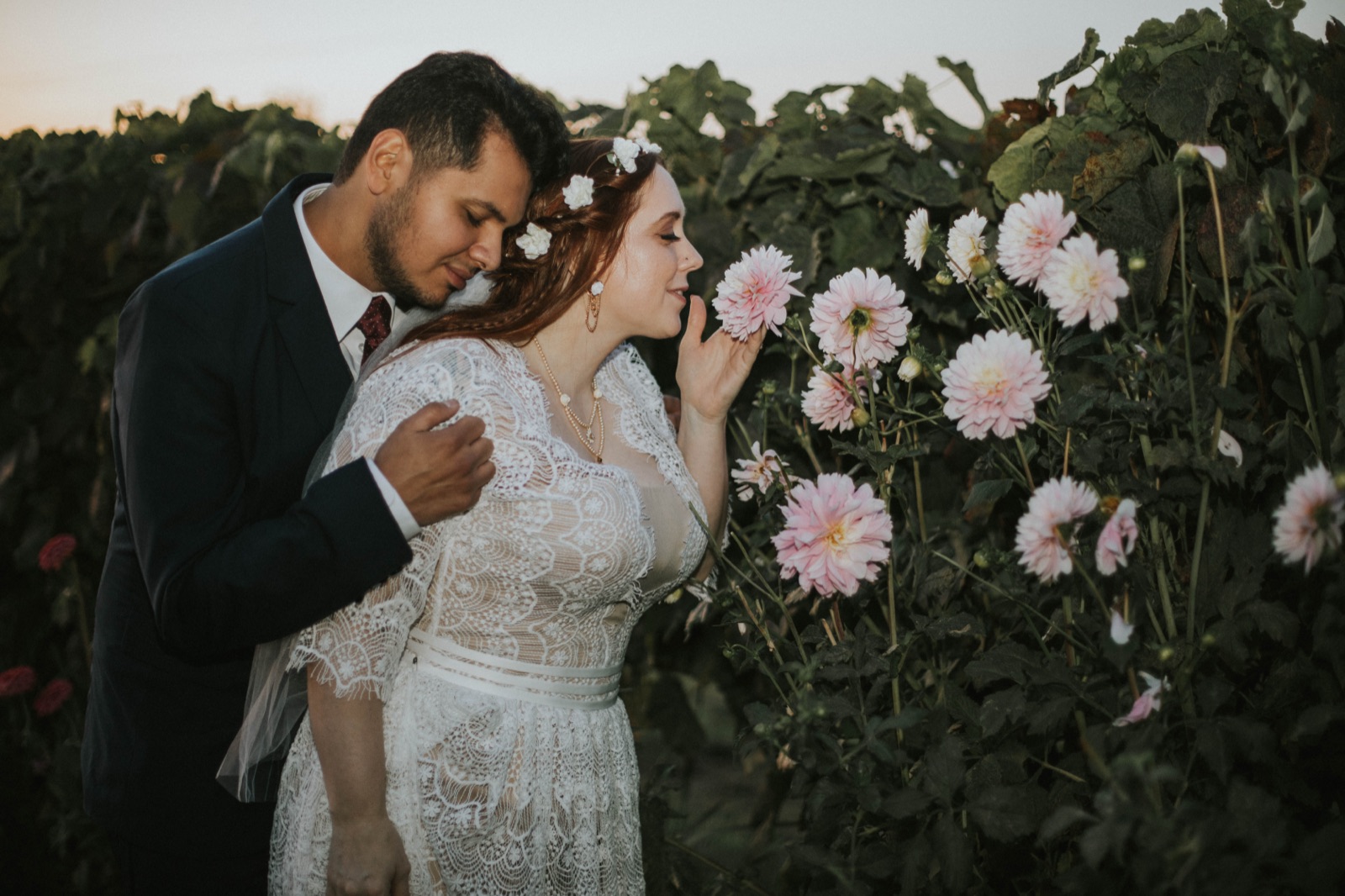 Groom embraces the bride from behind as she leans in to smell pink dahlias at dusk