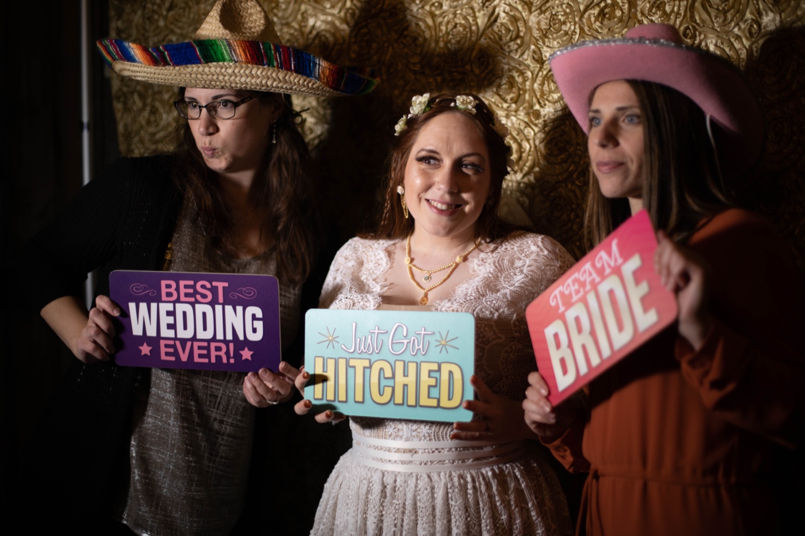 Bride and two guests hold Best Wedding Ever, Just Got Hitched, and Team Bride signs in front of a gold photo booth backdrop