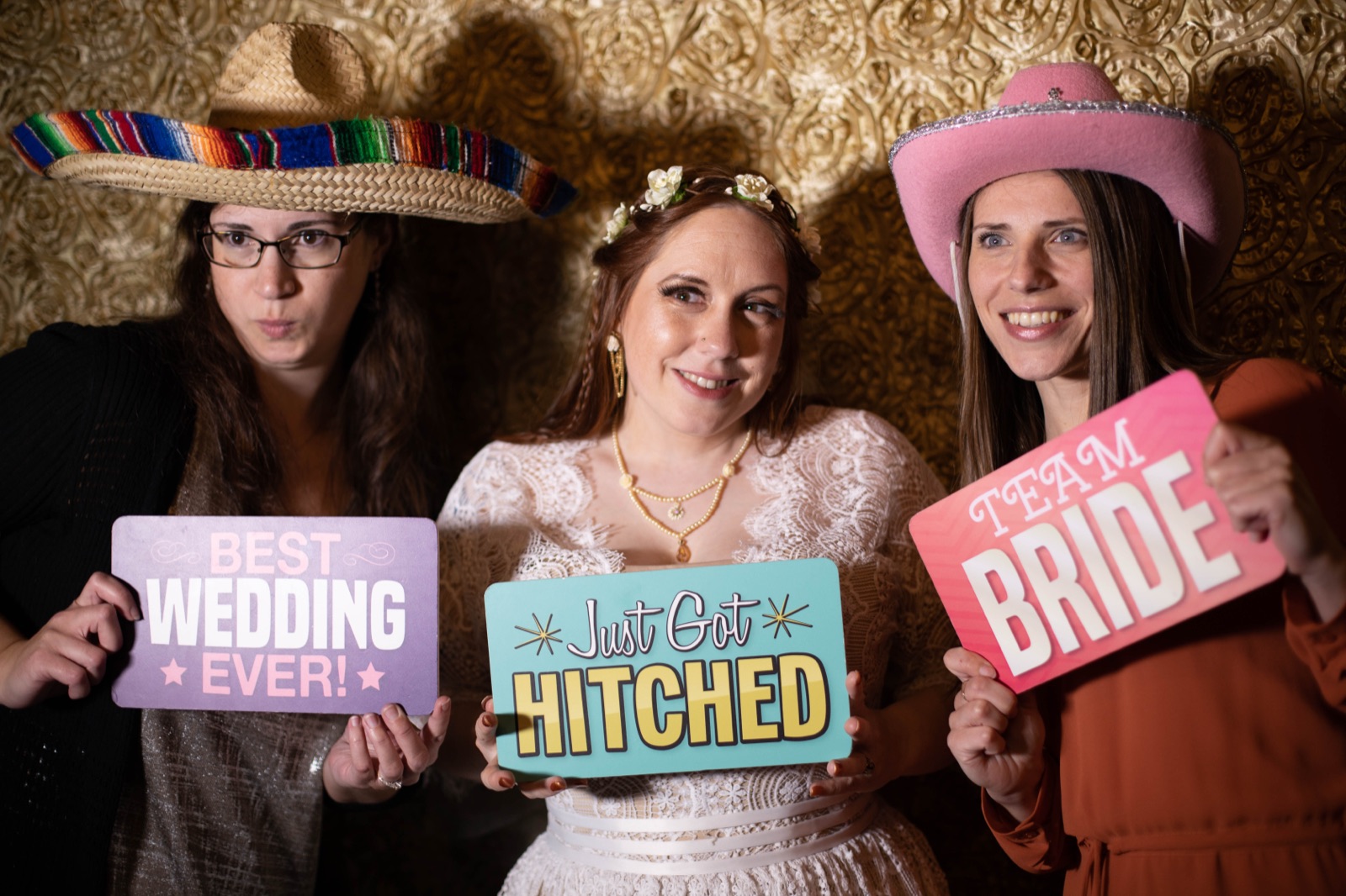 Three women in sombrero, flower crown, and pink cowboy hat hold wedding photo booth signs against a gold rosette backdrop