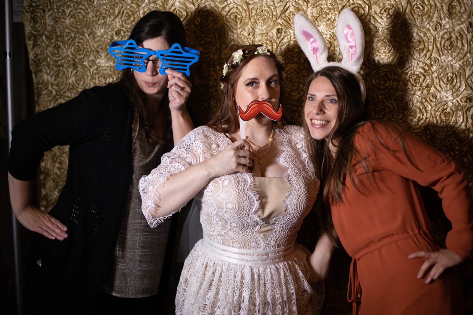 Bride with a mustache prop poses with two friends wearing star glasses and bunny ears in a photo booth