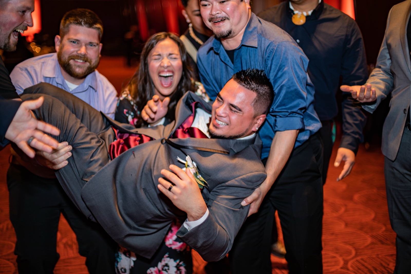 Laughing wedding guests lift the groom horizontally on a red-lit reception dance floor
