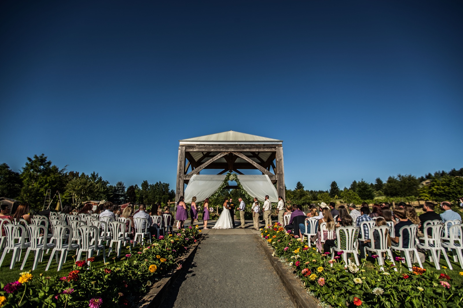 Outdoor wedding ceremony under a timber pavilion at the Oregon Garden, guests seated along a flower-lined aisle