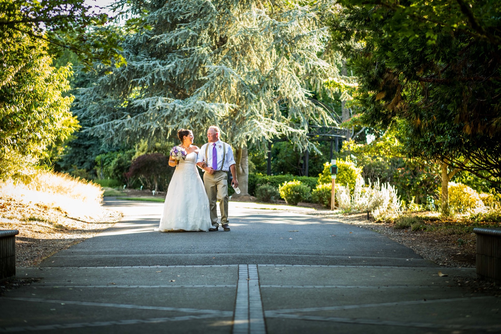 Bride and groom walk arm in arm down a tree-lined path at the Oregon Garden in late-day sun