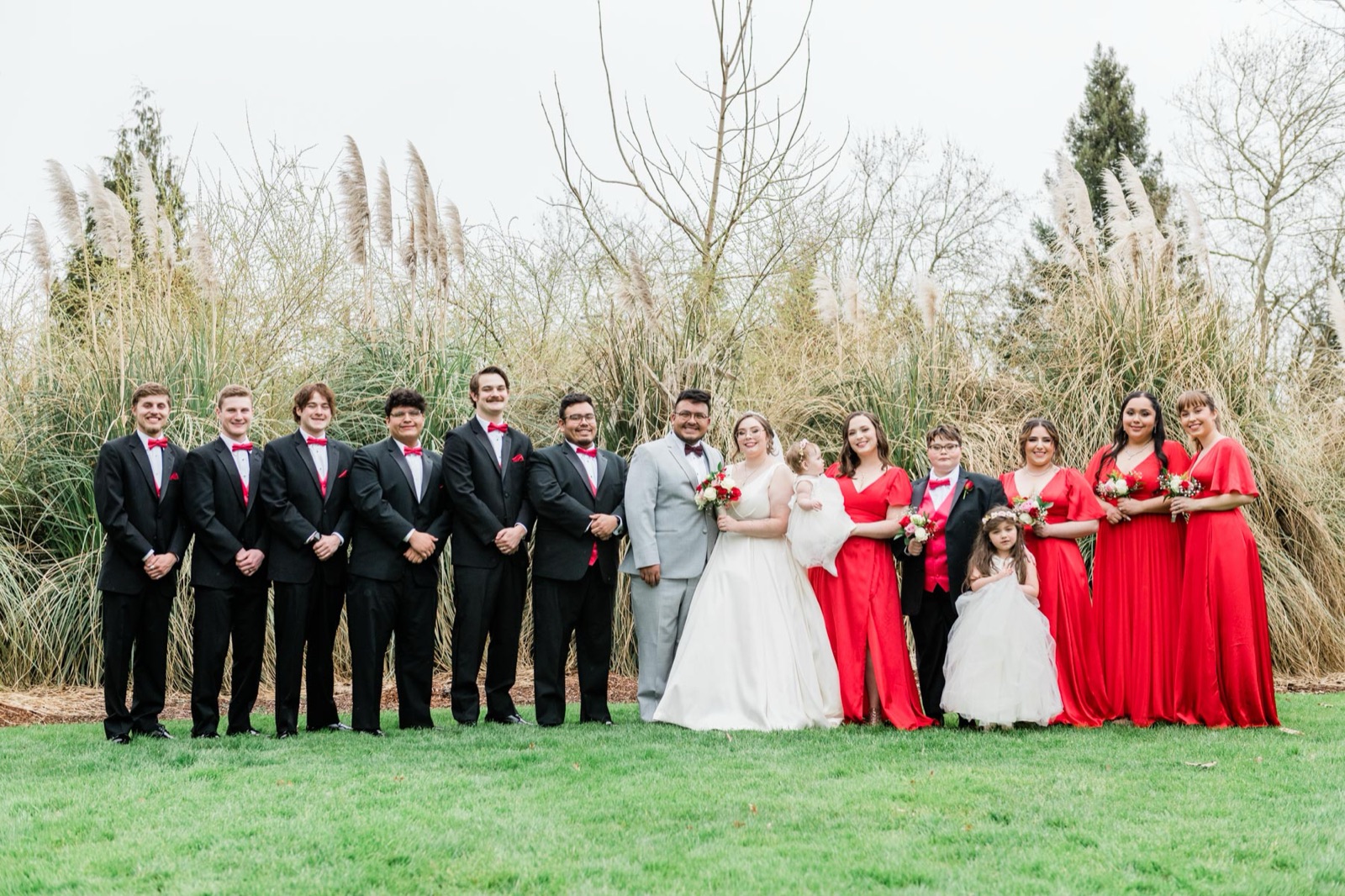 Full wedding party in black tuxes with red bow ties and red dresses posed on a lawn at Green Villa Barn in Independence, Oregon
