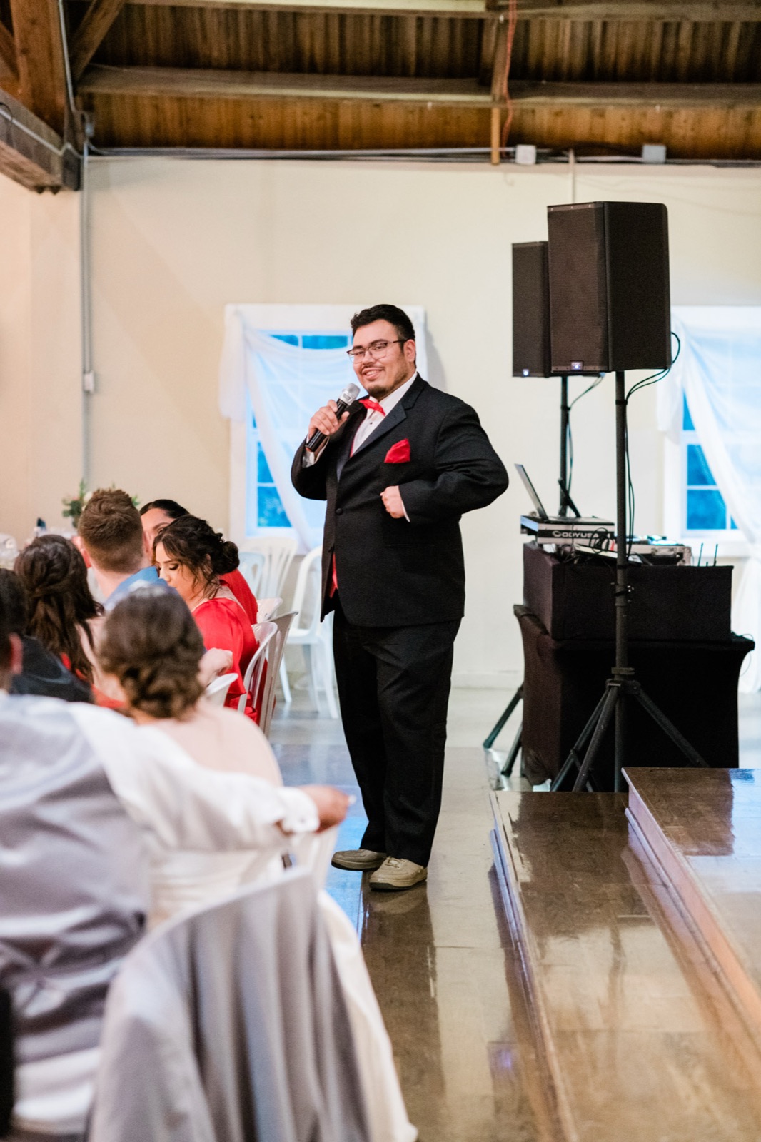 Emcee in a black suit with red pocket square speaks into a microphone beside a DJ speaker at Green Villa Barn in Independence, Oregon