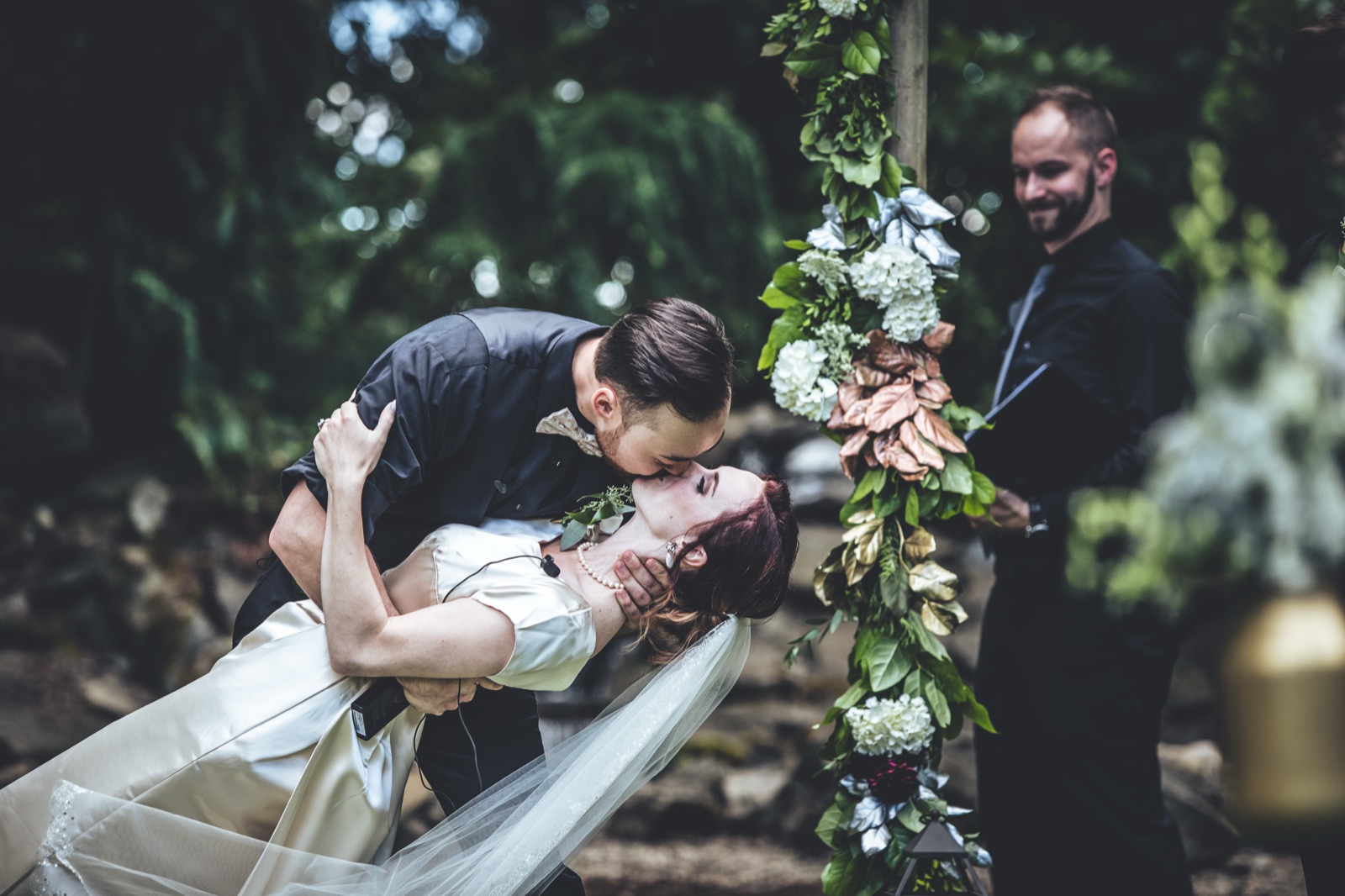 Groom dips and kisses the bride at an outdoor ceremony beside a flower-covered arch as the officiant smiles