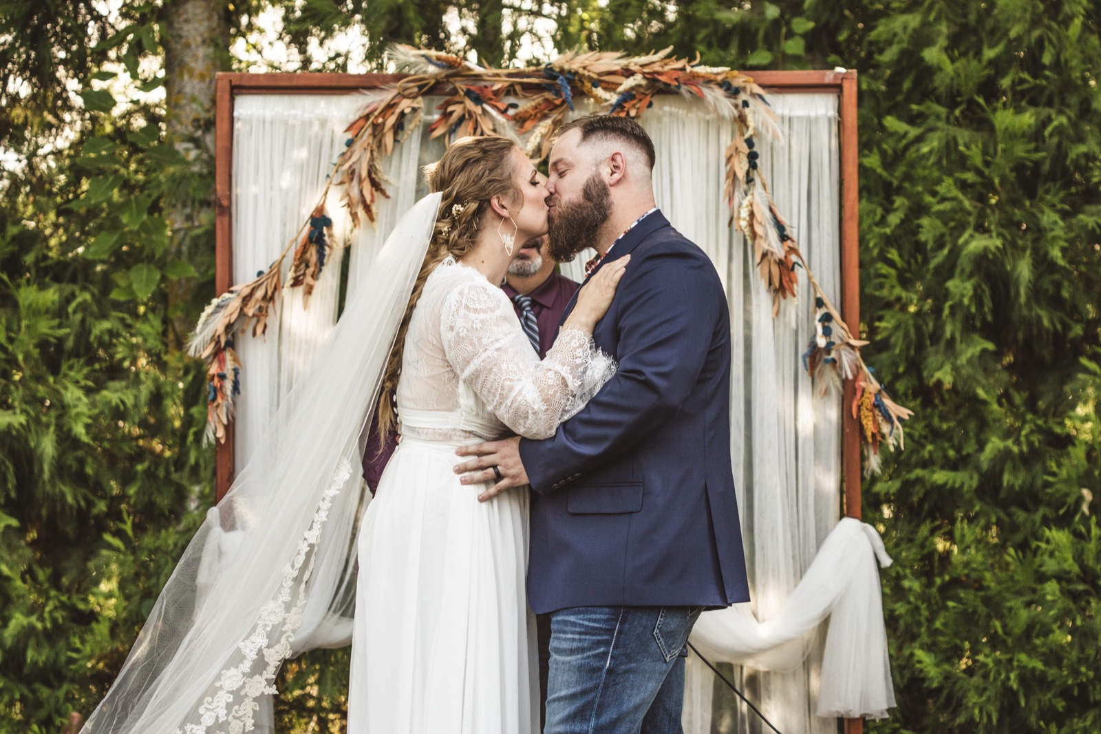 Bride and groom kiss at an outdoor ceremony in front of a wooden arch draped with fabric and dried florals