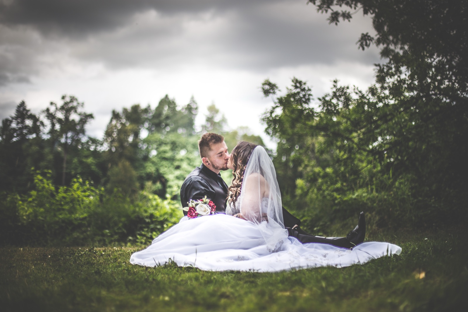 Bride and groom sit on the grass kissing, her dress and veil spread out, under a cloudy sky