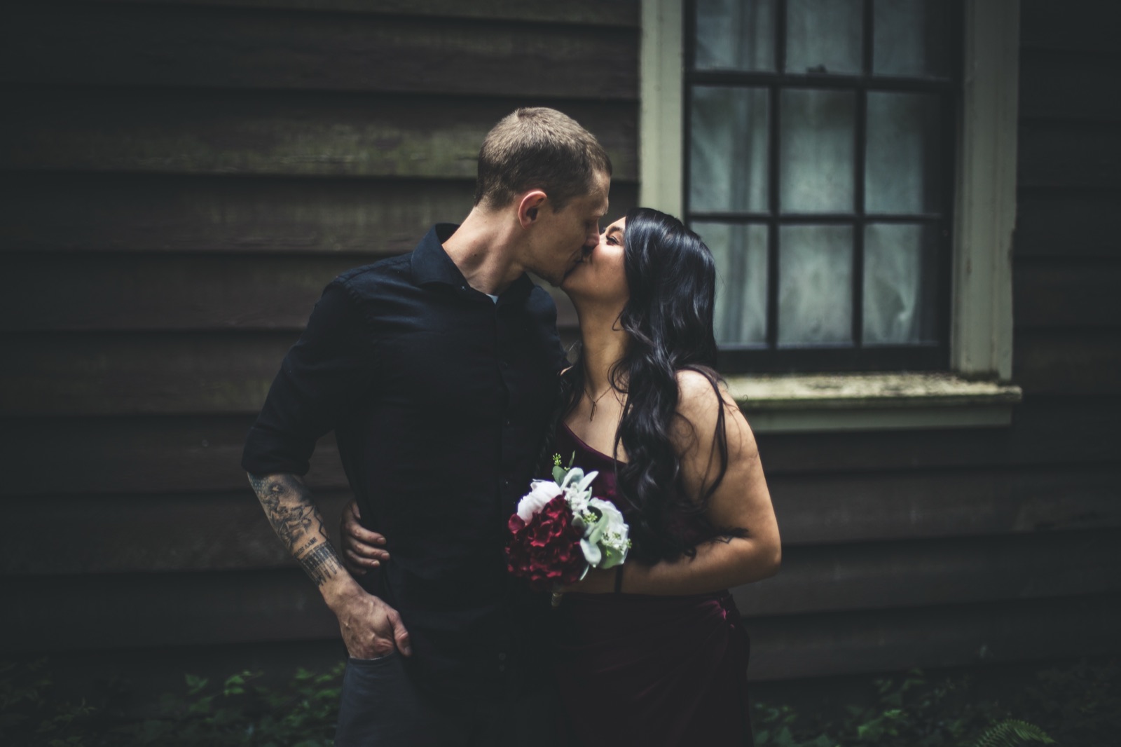 Couple kisses beside a dark wooden house, she in a burgundy dress holding a small red and white bouquet