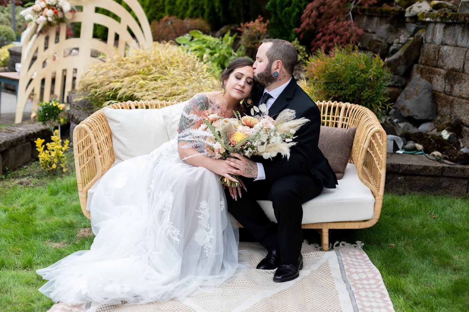 Groom kisses the bride on the forehead as they sit on a wicker sofa in a garden