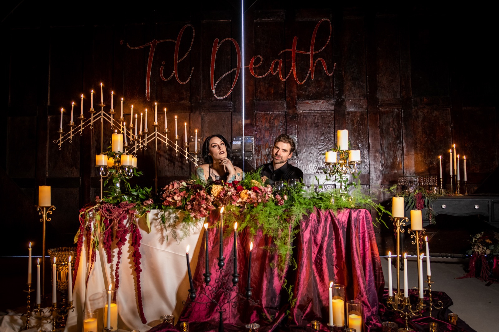 Couple seated at a candlelit sweetheart table with dark florals under a 'Til Death sign on a dark wood wall