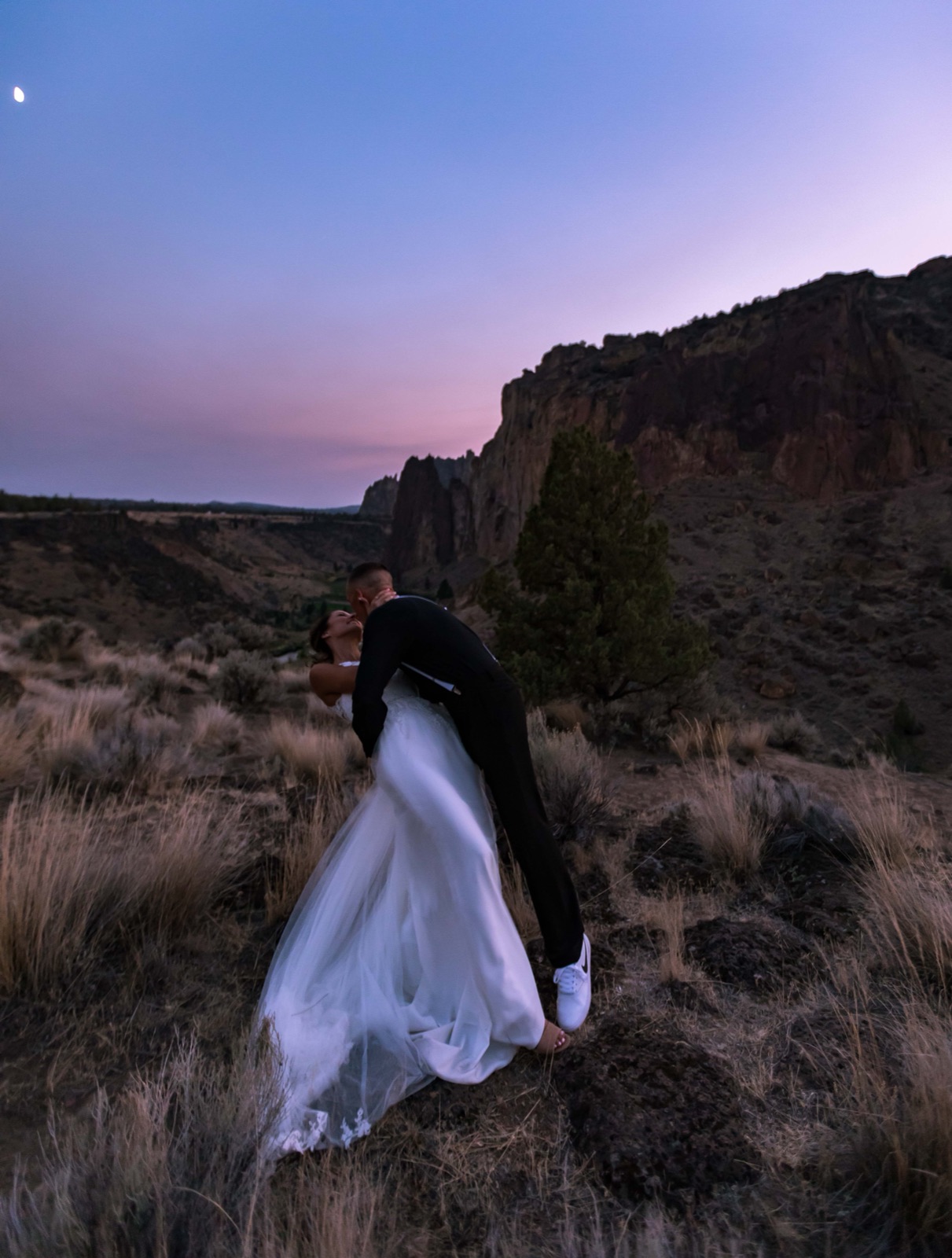 Groom dips and kisses bride in a white gown on a high-desert hillside at dusk, rock cliffs and moon behind them