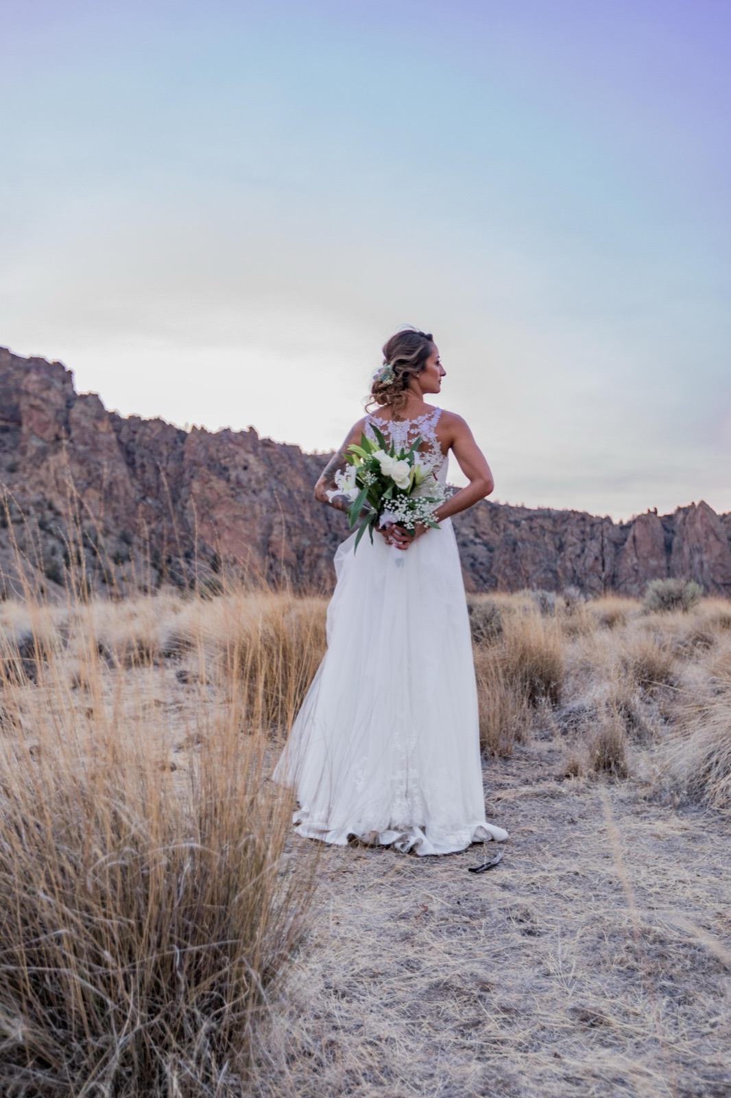 Bride seen from behind holding her bouquet at her back, looking toward canyon cliffs at dusk