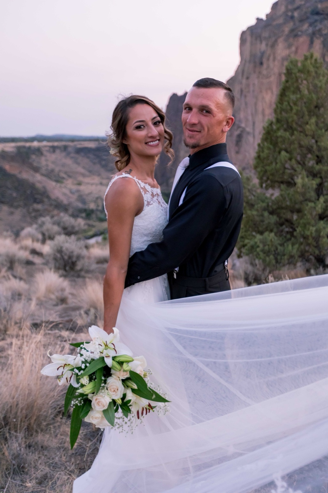 Bride and groom smiling at the camera above a canyon, her veil sweeping across the foreground
