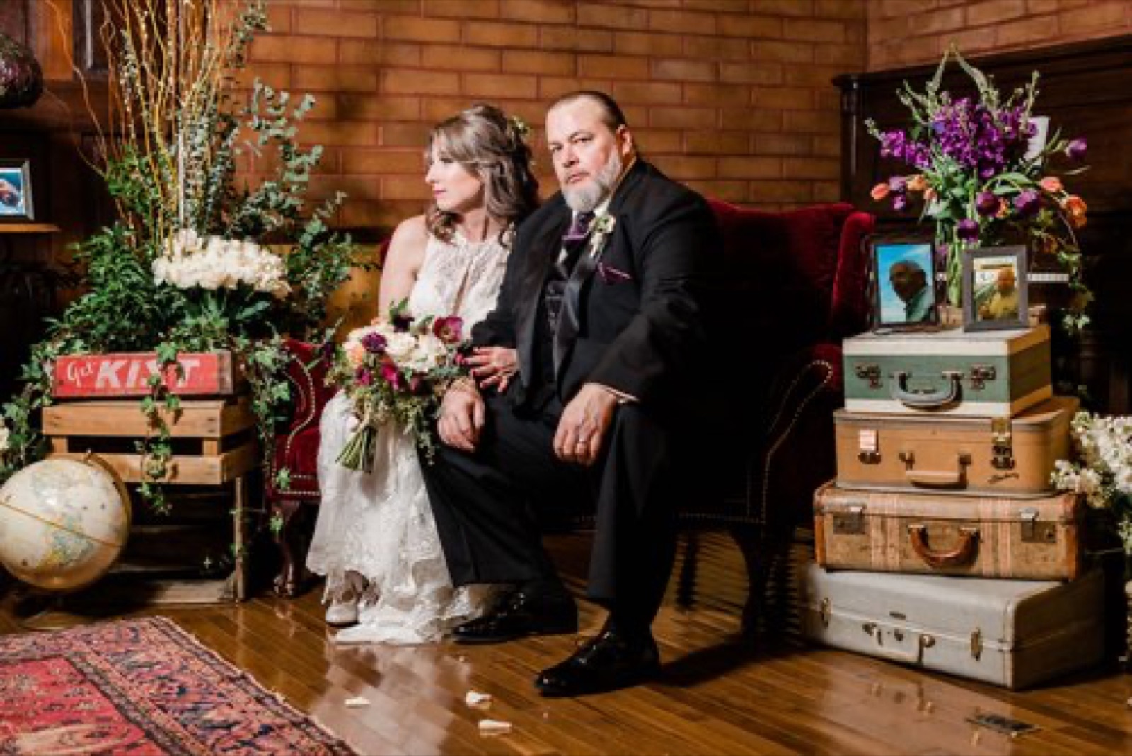 Bride and groom seated on a red velvet settee among vintage suitcases and flowers