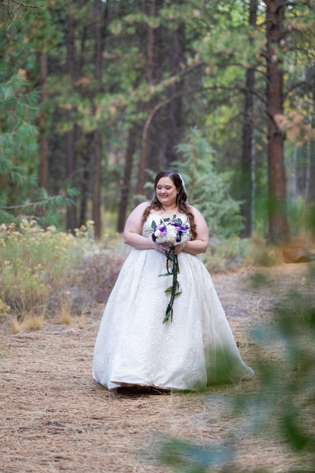 Bride in a lace ball gown with a purple and white bouquet on a pine-forest path