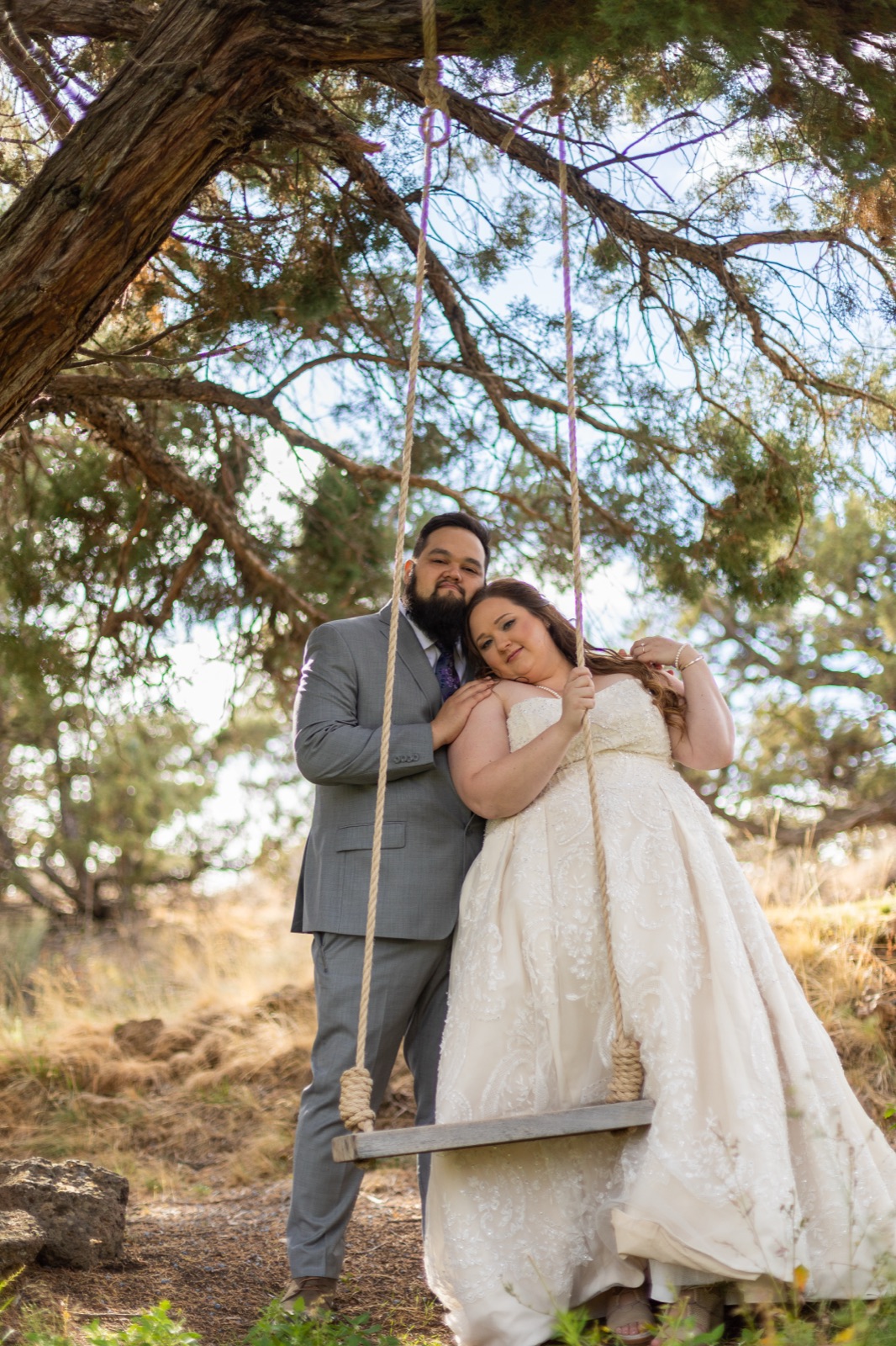 Groom stands beside bride at a rope tree swing under a juniper, both looking at the camera