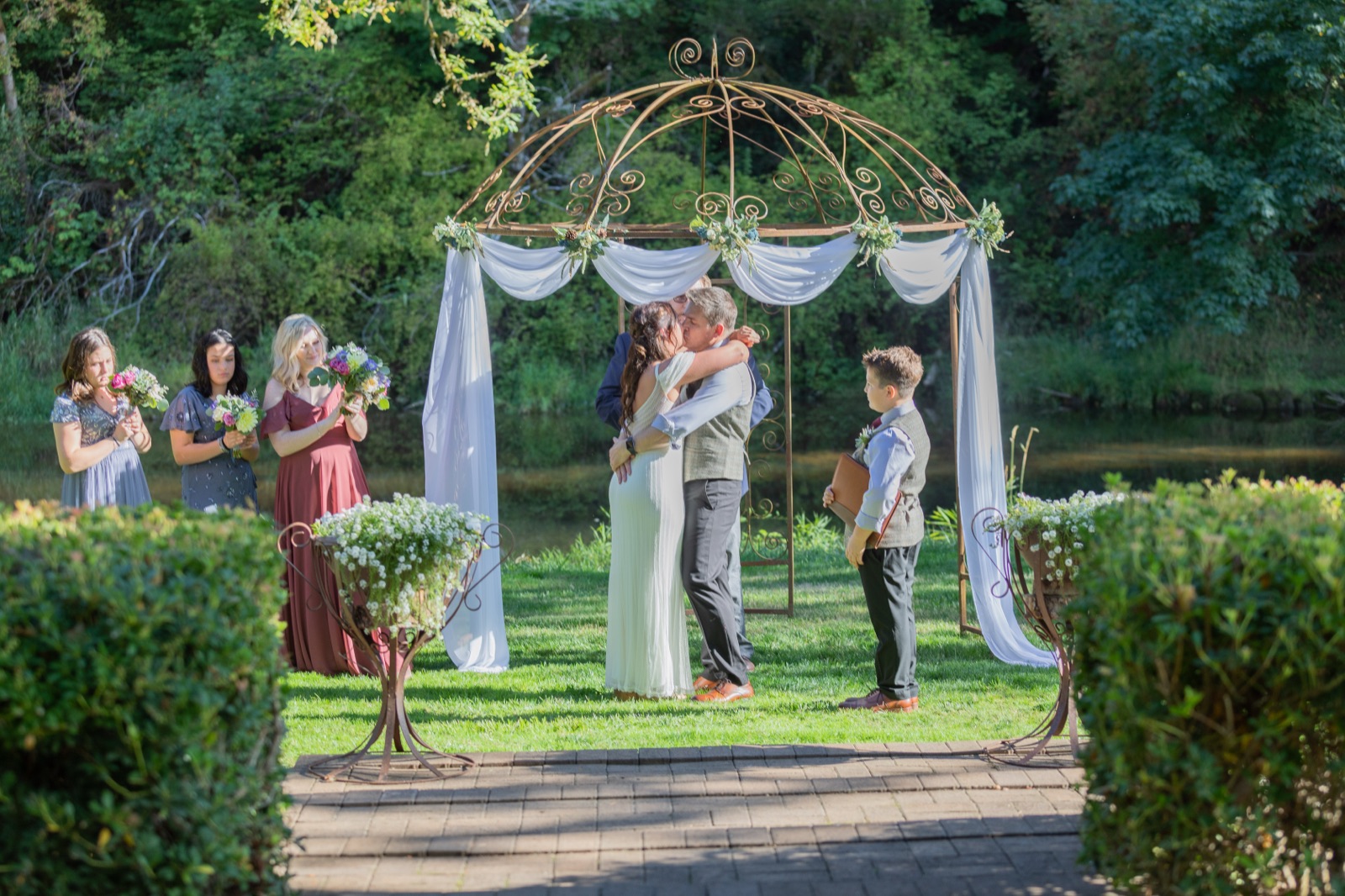 Couple kisses under a wrought-iron gazebo draped in white fabric beside a pond as bridesmaids and a boy look on