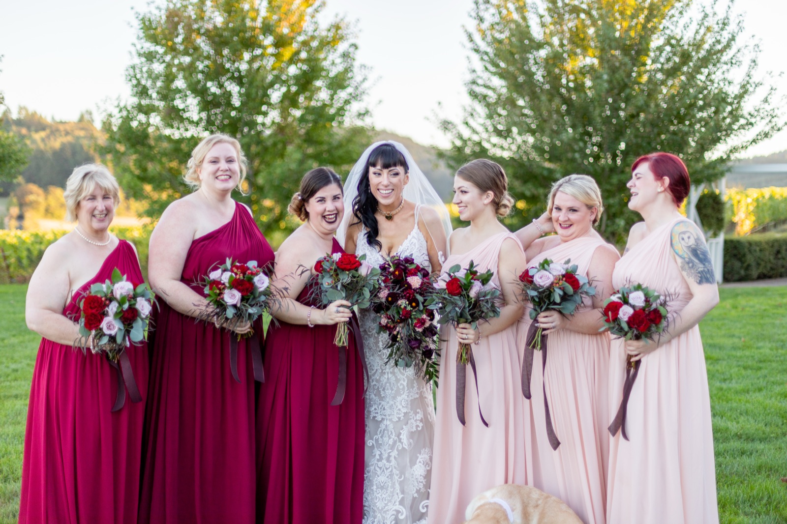 Bride laughs with six bridesmaids in red and blush gowns holding rose bouquets on a vineyard lawn
