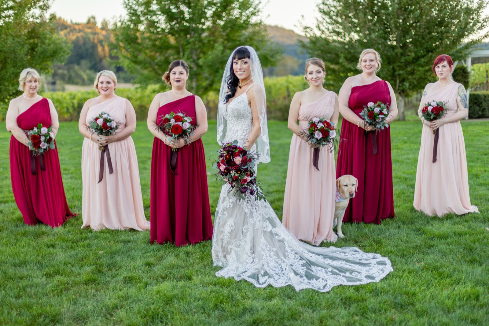 Bride with lace train stands with bridesmaids in red and blush dresses and a yellow lab on green grass