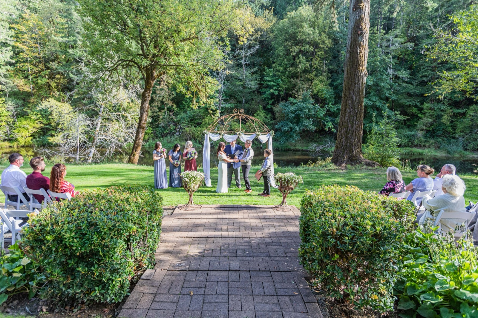 Small outdoor wedding ceremony under a metal arch beside a pond, guests seated on white chairs