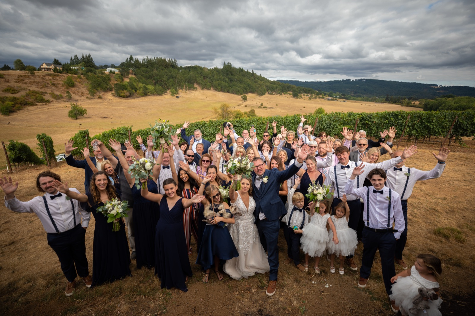 Bride, groom, and all their guests raise their hands and cheer on a hillside beside vineyard rows