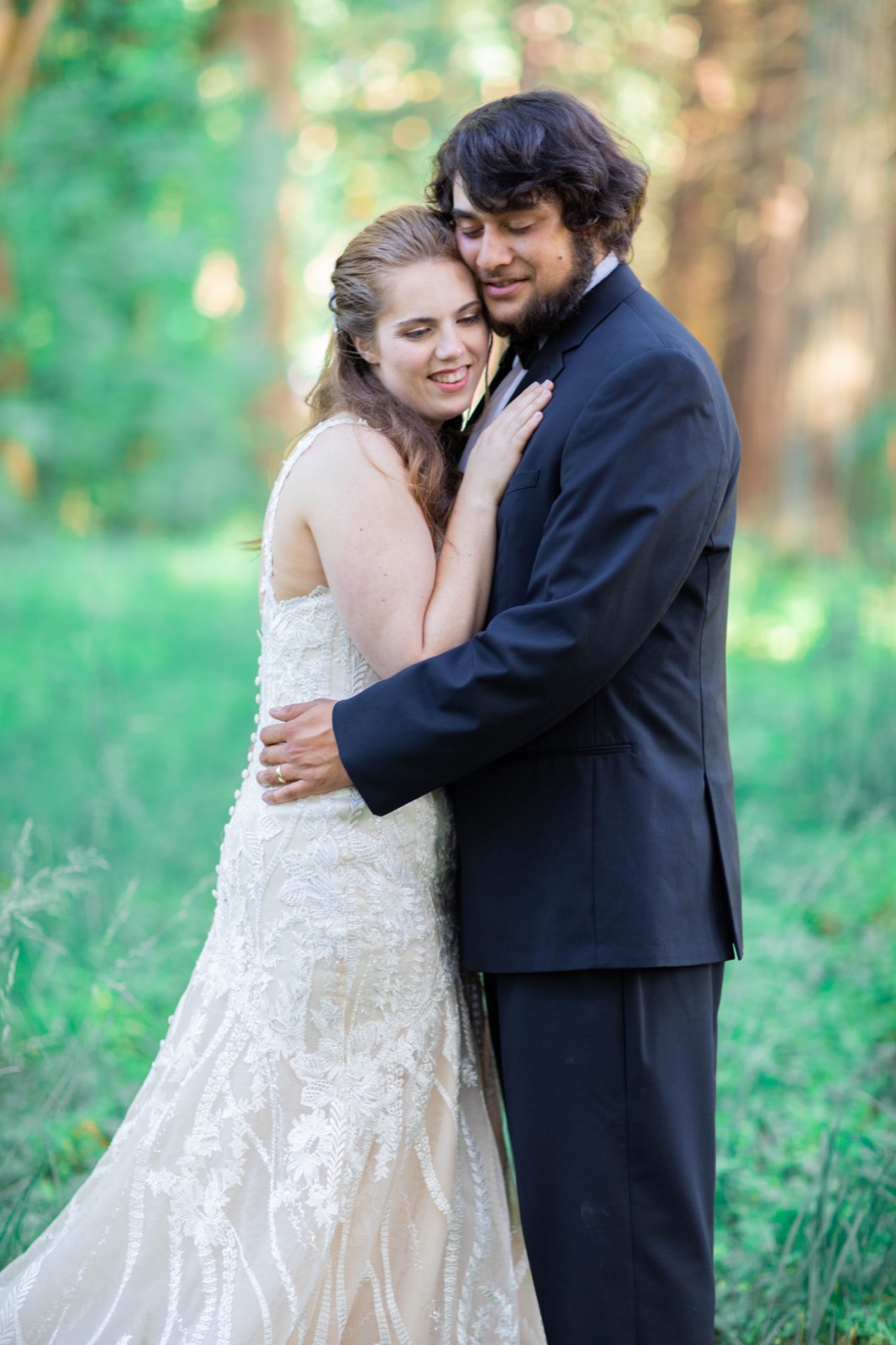 Bride rests her head against the groom's cheek as they embrace in a sunlit forest clearing