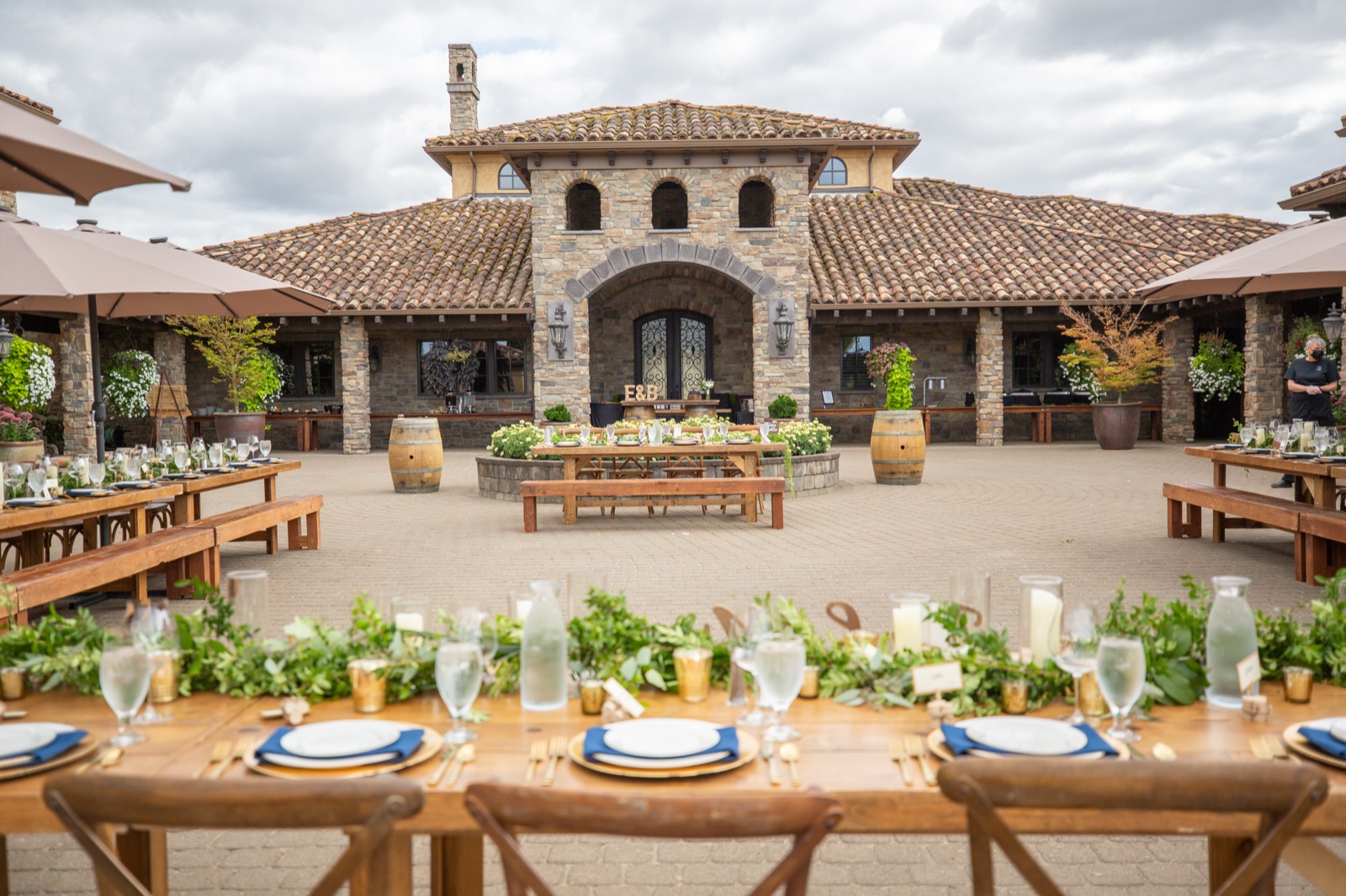 Farm tables set with greenery, candles, and navy napkins in a stone winery courtyard