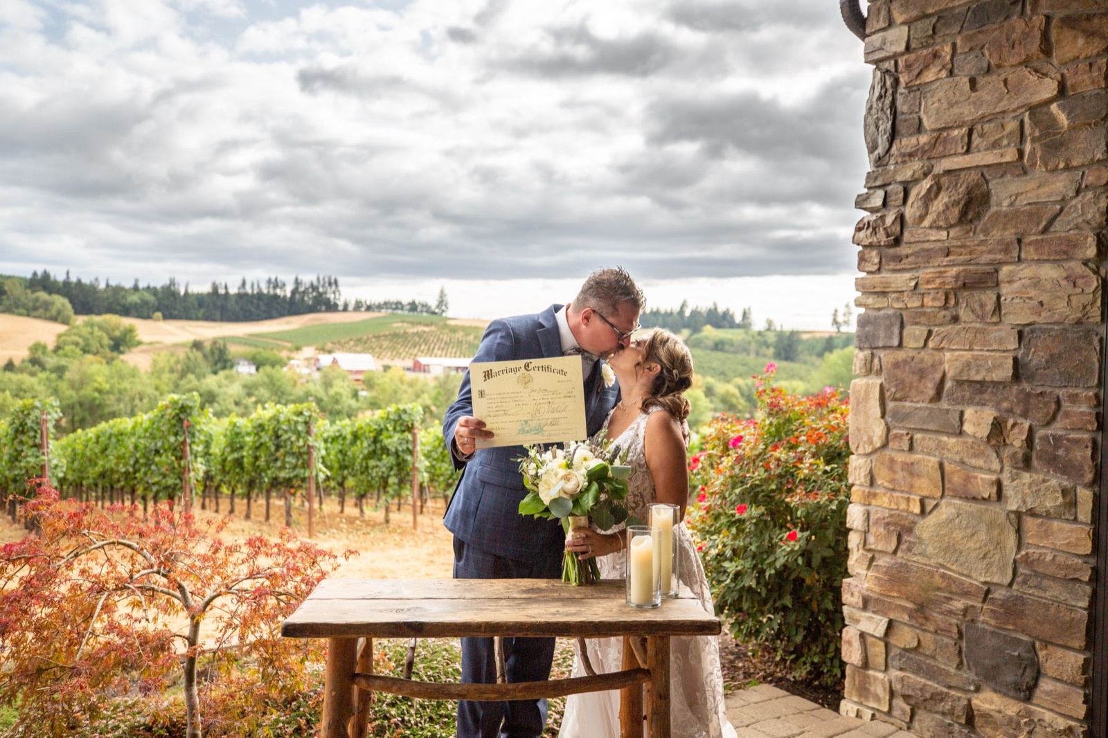 Bride and groom kiss while holding their marriage certificate on a stone patio overlooking a vineyard