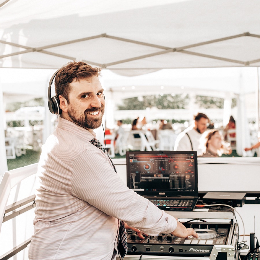 Allen Carter in headphones smiling back from his DJ decks under a tent at an outdoor wedding