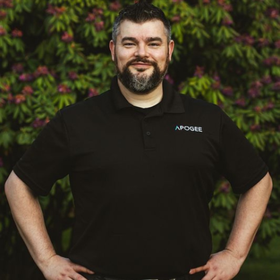 Jack Harris smiling in a black Apogee polo in front of flowering bushes