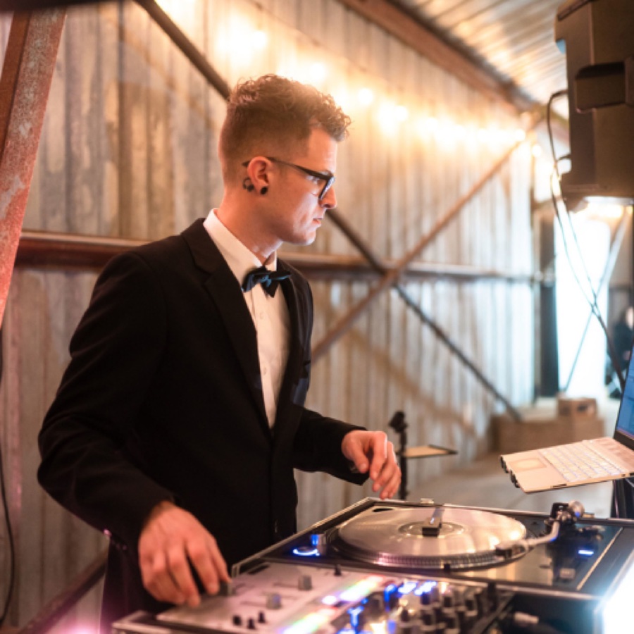 Kyle Epps in a tuxedo and bow tie mixing on a turntable under string lights
