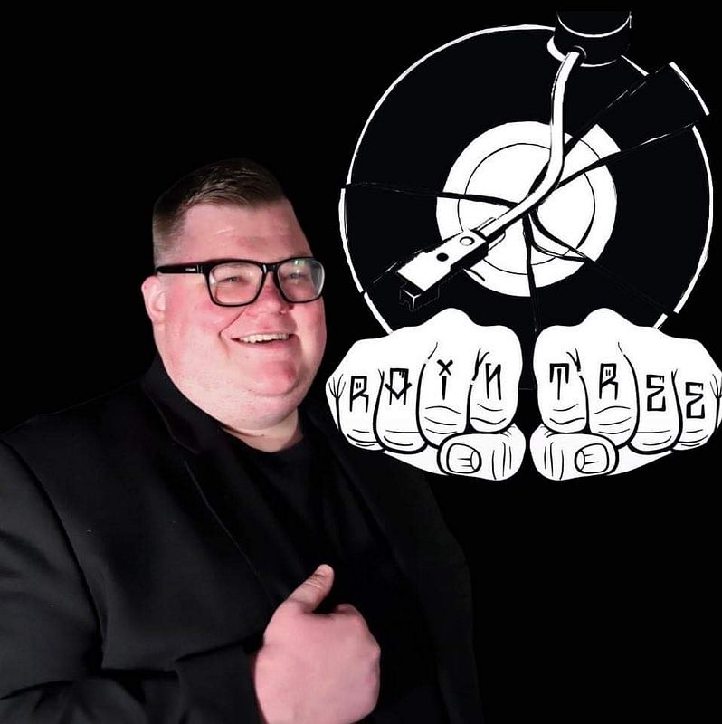 Rian Simpson, DJ Raintree, smiling in a black jacket beside his Raintree vinyl-record logo