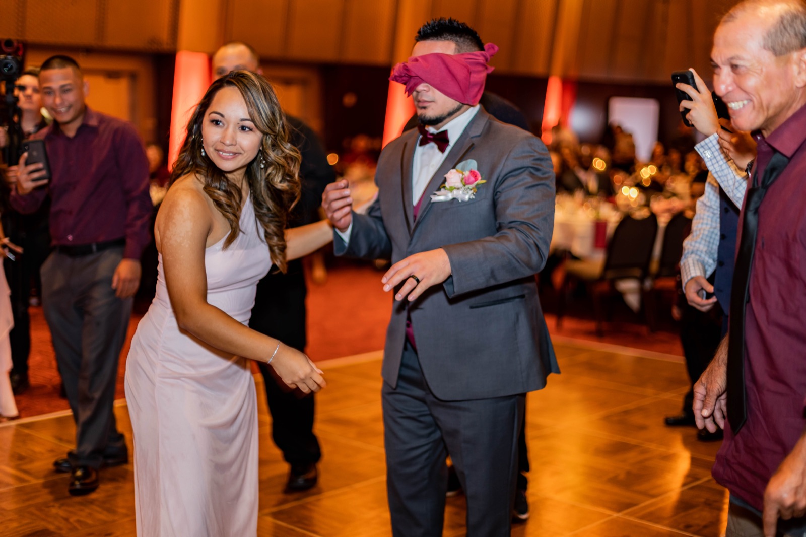 Blindfolded groom in a gray suit guided across the dance floor as laughing guests film on phones
