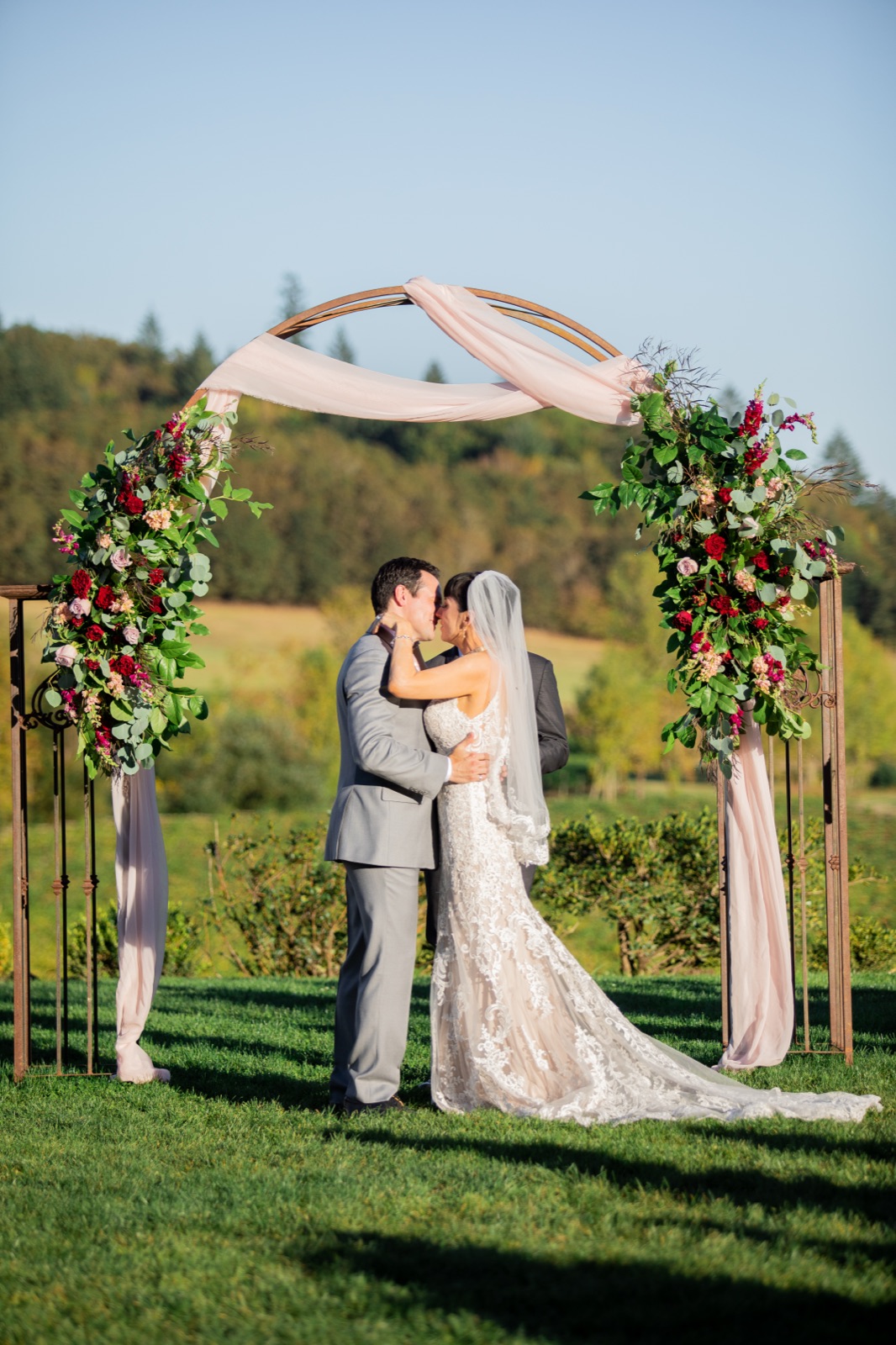 Bride and groom kiss under a round metal arch with greenery and blush drapery at Zenith Vineyard